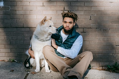 A man with a beard and dreadlocks sits on the ground with a white dog.