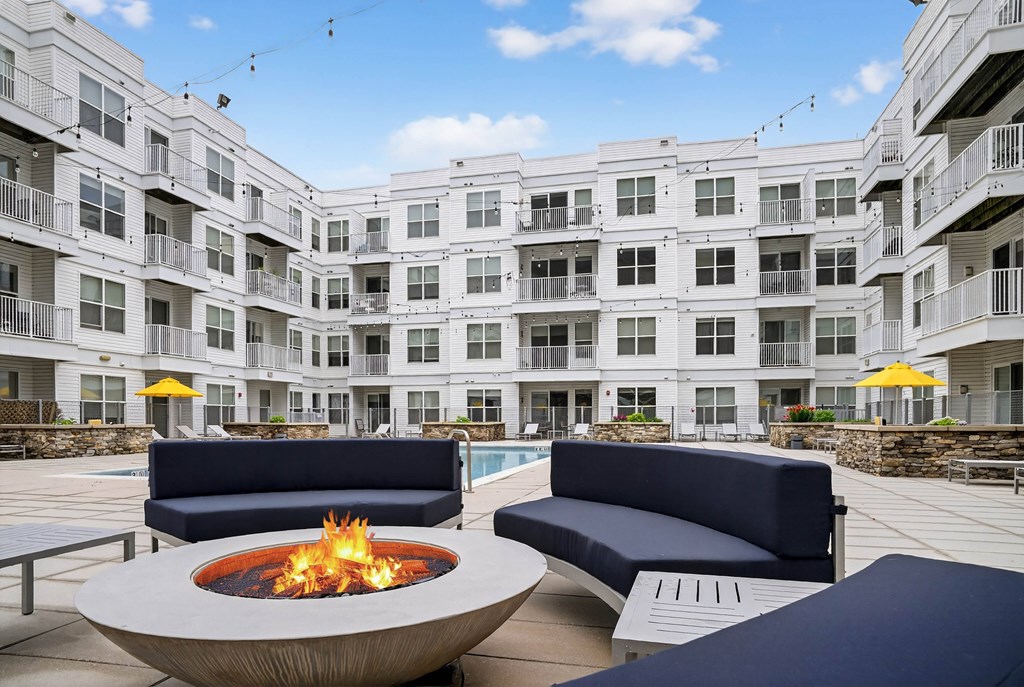 A fire pit sits in the middle of a patio with blue couches and a pool in the background.