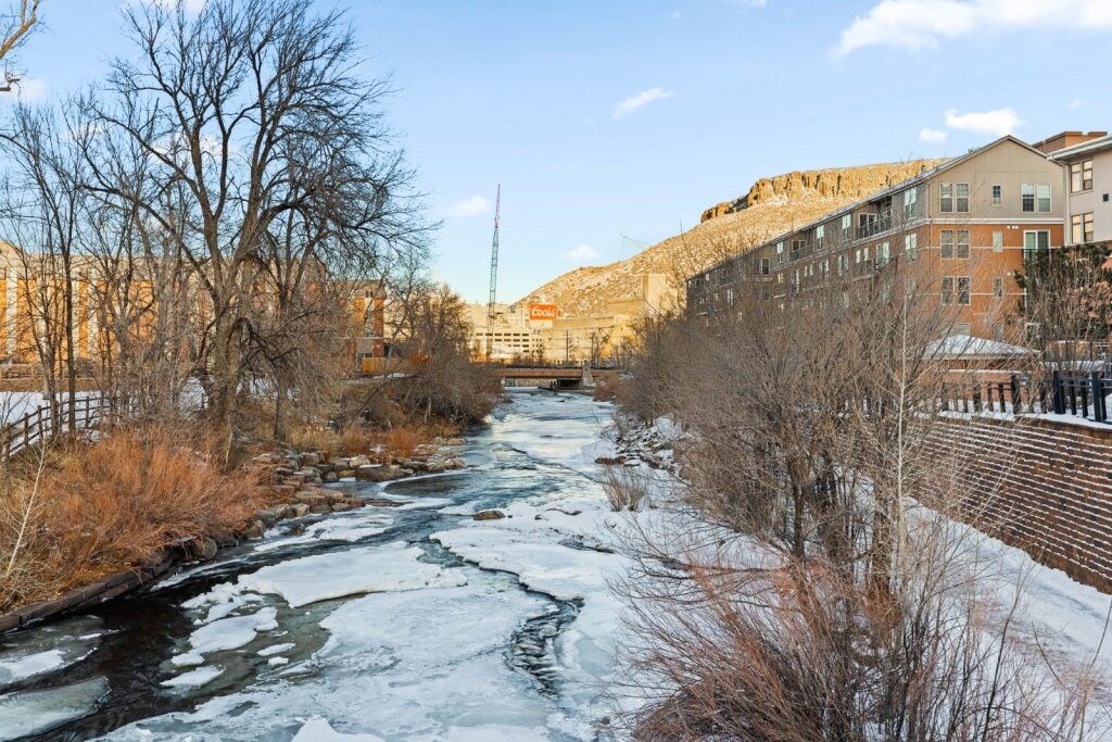 A river flows through a snowy landscape with buildings on the side at The Avenue Lofts Golden Apartments, Colorado, 80401