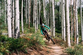 A person in a green jacket and white helmet is riding a bicycle on a trail in a forest of birch trees at The Avenue Lofts Golden Apartments, Golden, CO