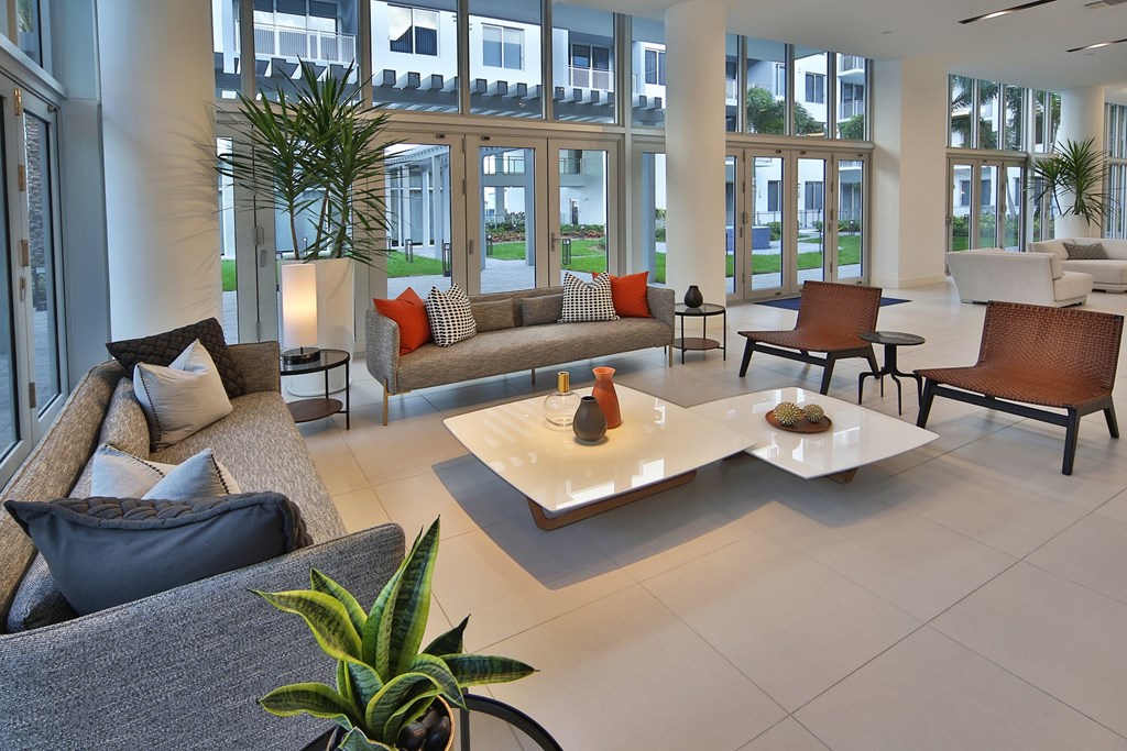 Clubhouse with open layout with large windows that bring in natural light and offer a view of the outdoor space. The seating area features comfortable, modern furniture, including gray sofas with accent pillows, stylish brown woven chairs, and sleek, minimalistic tables. Potted plants and decorative vases add a touch of greenery and warmth, enhancing the inviting atmosphere of the space.