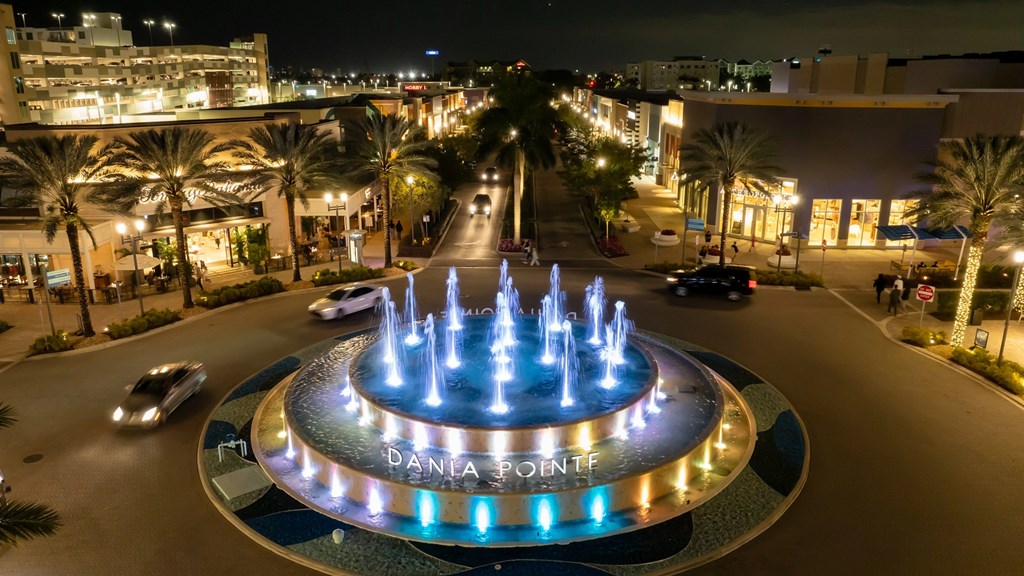 A lit-up water fountain at night in the middle of a plaza and roundabout with the name Dania Pointe on it.