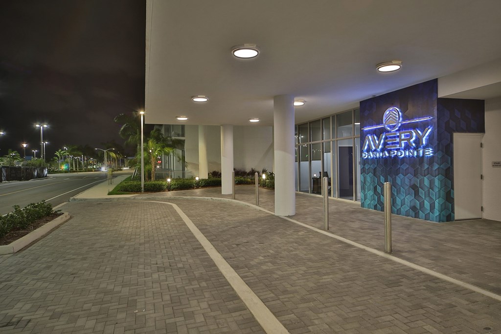 The entrance to this apartment building features a modern, well-lit design with sleek columns, a paved walkway, and an illuminated sign that reads "Avery Danna Pointe" in blue neon. The surroundings are lush with greenery, and the area has a clean, contemporary look, illuminated by overhead lighting.