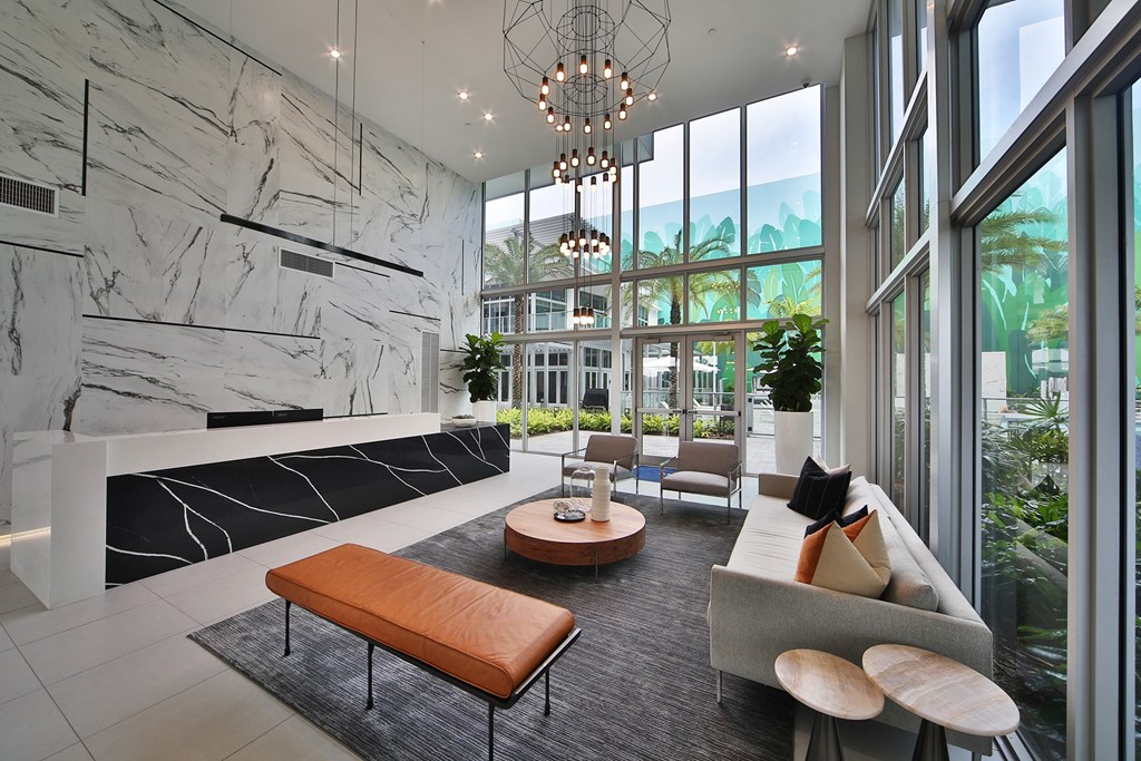 Lobby with a stunning marble front desk with a black and white design, seamlessly integrated into the space. The modern aesthetic is complemented by sleek marble walls, large windows allowing abundant natural light, and a stylish seating area with a leather bench and contemporary furniture. Potted plants and unique lighting fixtures further elevate the sophisticated setting.