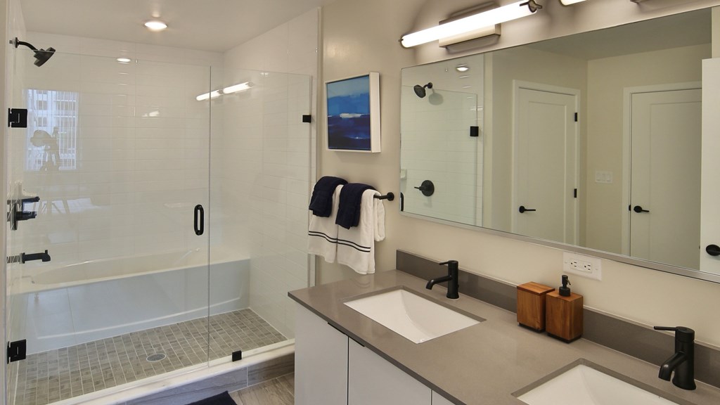 Modern residence bathroom featuring a sleek, minimalist design with a spacious separated shower and tub combo enclosed by clear glass panels. The vanity area includes dual sinks with matte black faucets, a large mirror, and a clean countertop with white cabinets undearneath. The room is illuminated with bright, contemporary lighting.