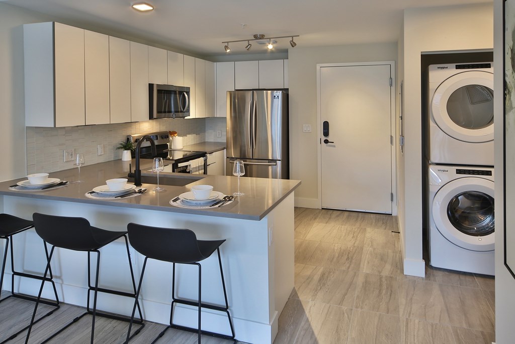 Residential kitchen featuring bar stool seating, black fixtures in the kitchen, white cabinets, quartz countertops, stainless steel appliances with barn door fridge. Off to the side a full-sized  stackable washer and dryer.