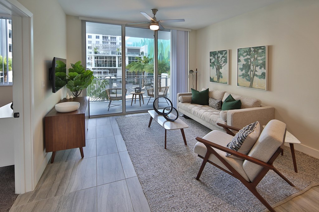 A modern apartment living room featuring neutral tones, mid-century furniture, and floor-to-ceiling triple-panel balcony doors that overlook the resort-style pool. The space includes a beige sofa, accent chairs, marble-top coffee table, and nature-inspired artwork, creating a stylish yet comfortable atmosphere.