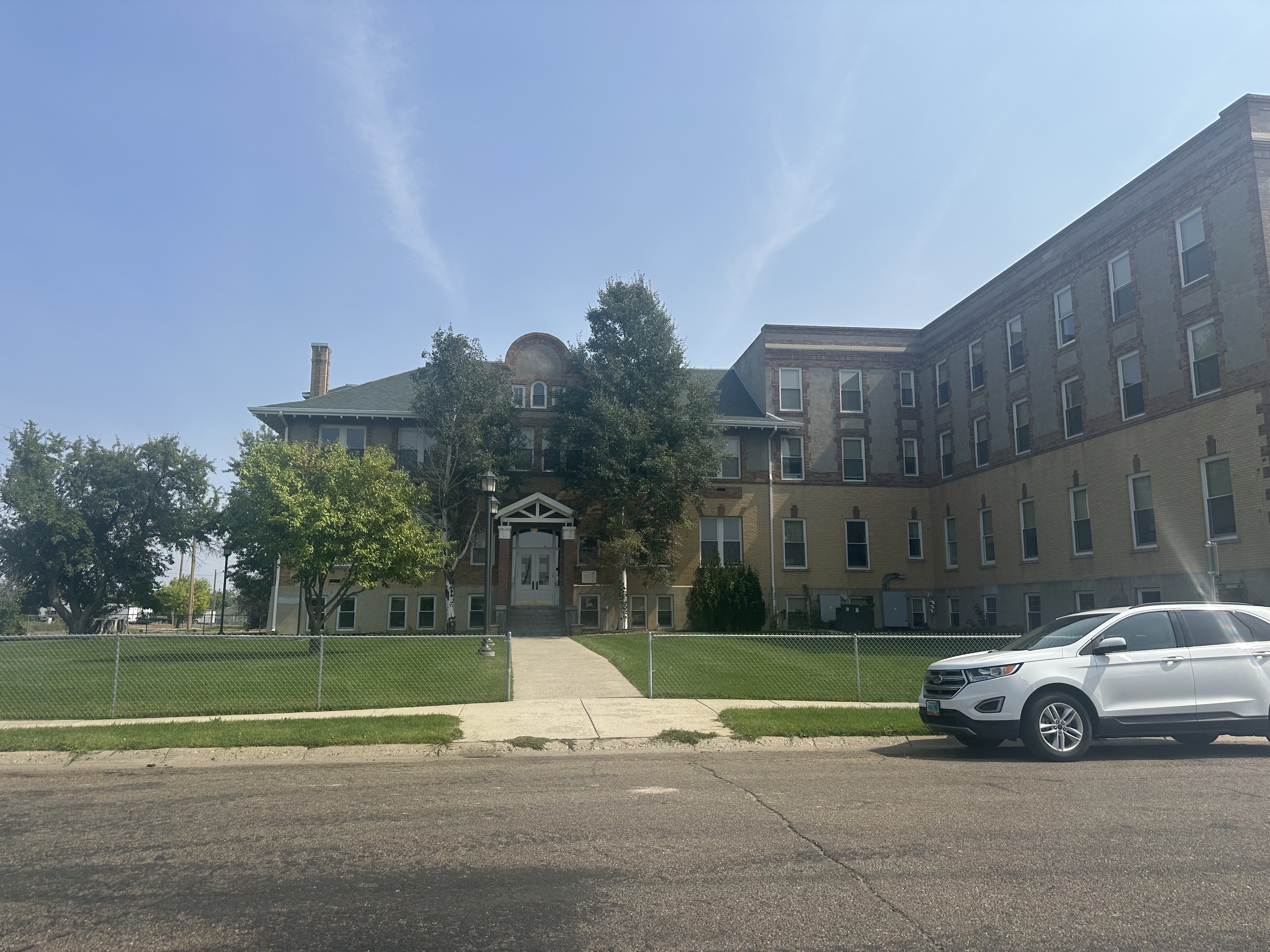 A white car is parked in front of The Cornerstne Apartments with a fence and trees.