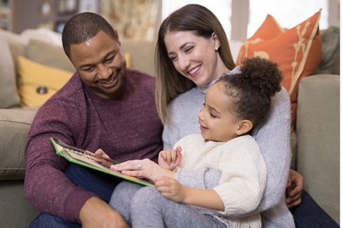 A family of three is sitting on a couch, reading a book together.