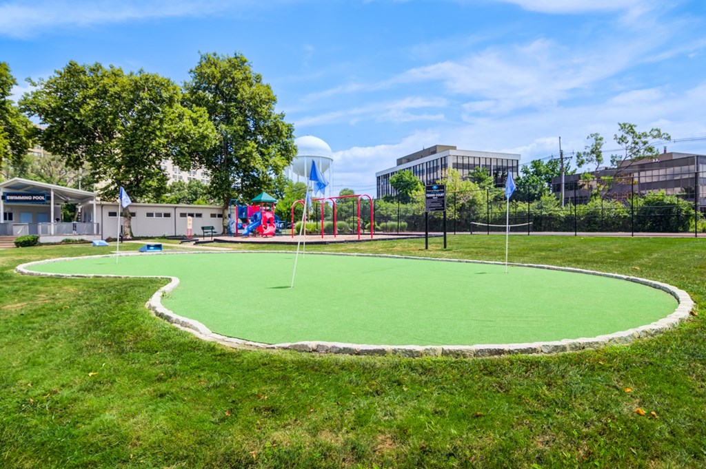A green artificial turf in the foreground with a playground and buildings in the background.