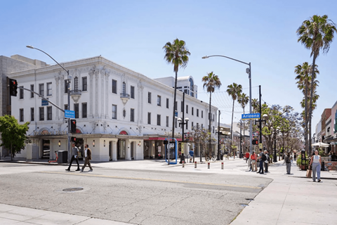 A street scene with a white building, palm trees, and people walking.