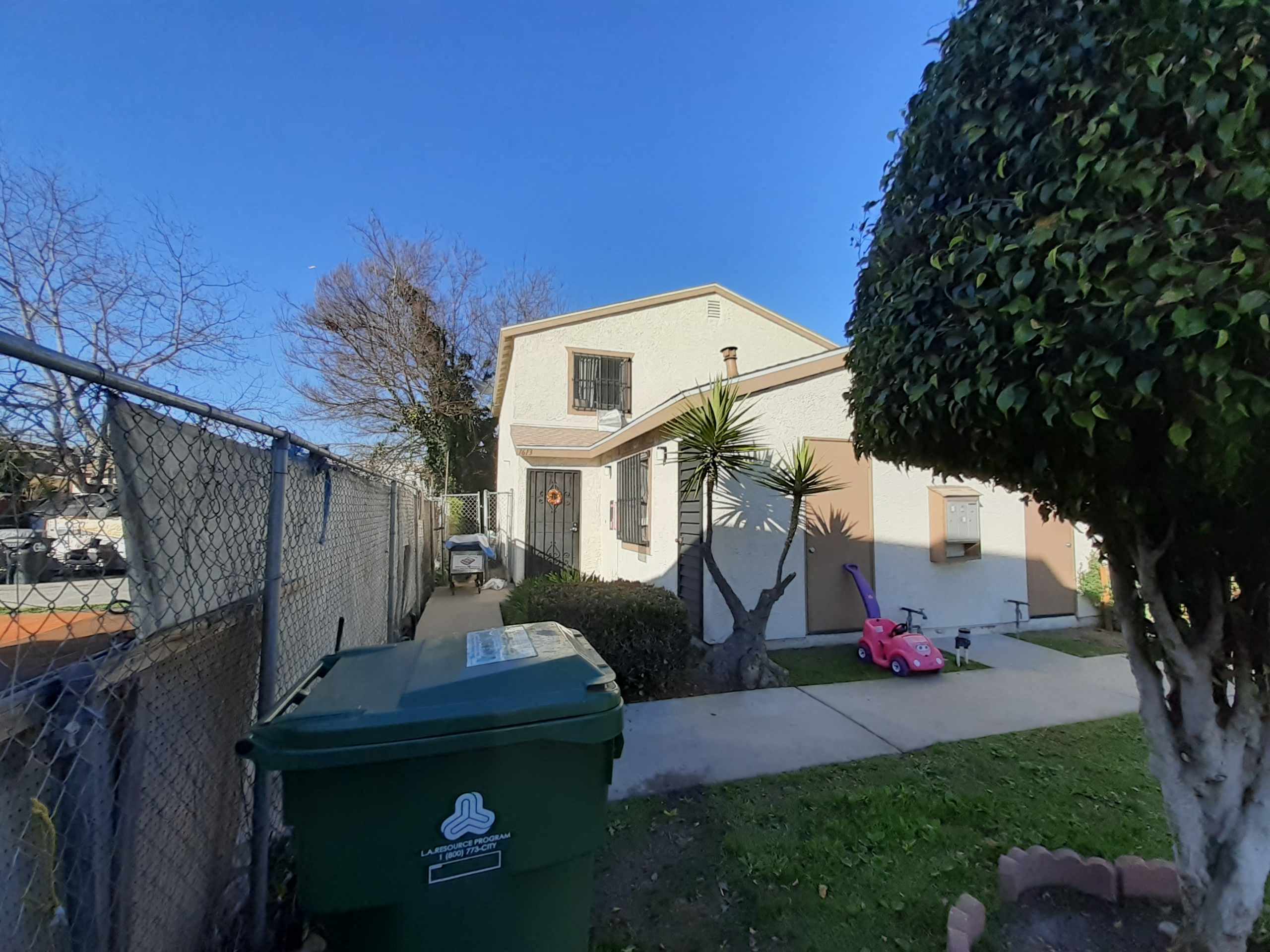 A green trash bin with a logo on it is in the foreground of a residential street.
