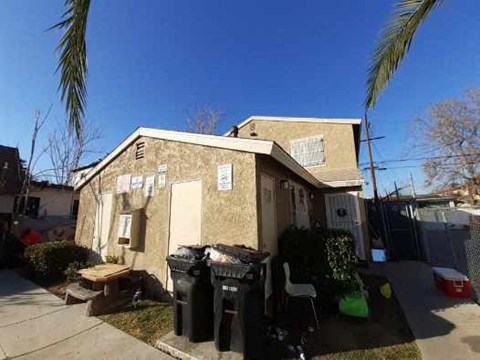 A house with a brown wall and a white door is surrounded by a fence and has a green lawn.