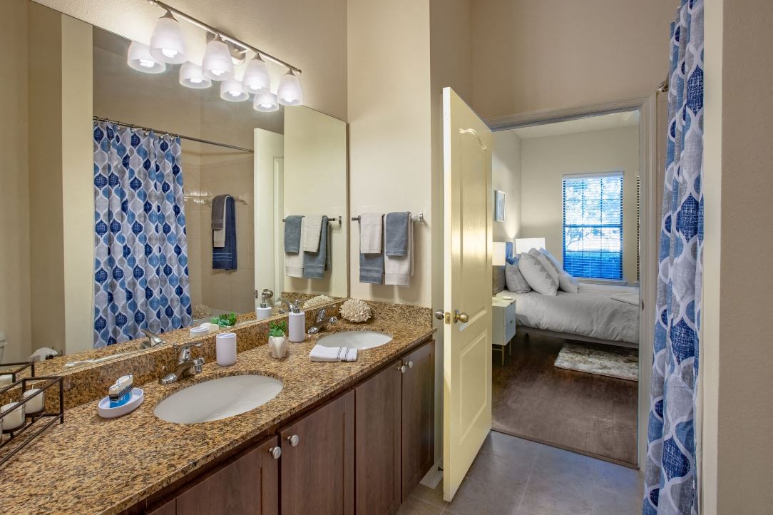 Bathroom with double vanity sinks pictured. Cabinets are dark brown with granite counter tops. Bathroom entry located within one of the bedroom at The Bartram Apartments in Gainesville, FL 32608