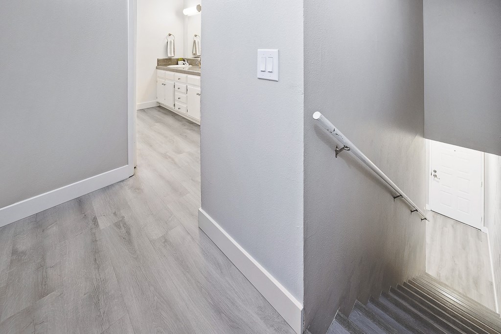 an empty hallway with a staircase and a bathroom with white cabinets and wood flooring