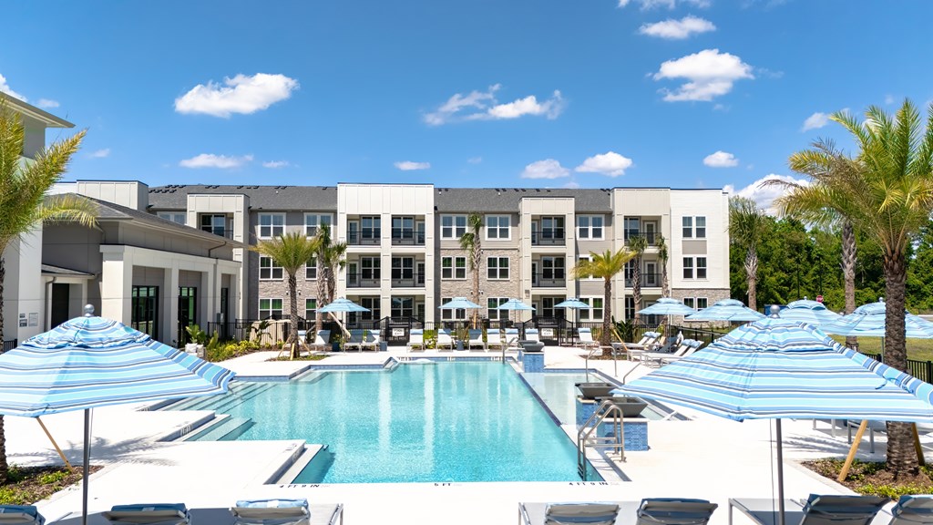 A large swimming pool surrounded by lounge chairs and umbrellas in front of a multi-story building.
