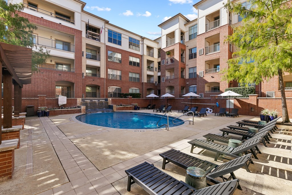 A large outdoor pool area with sun loungers and umbrellas in front of apartment buildings.
