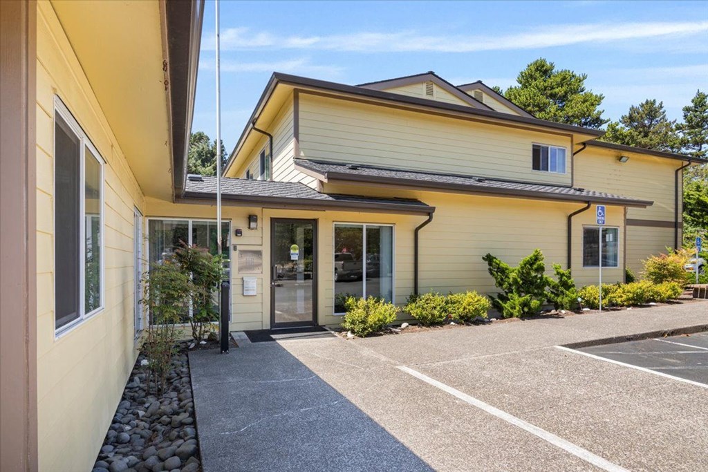 A yellow building with a grey roof and a parking lot in front.