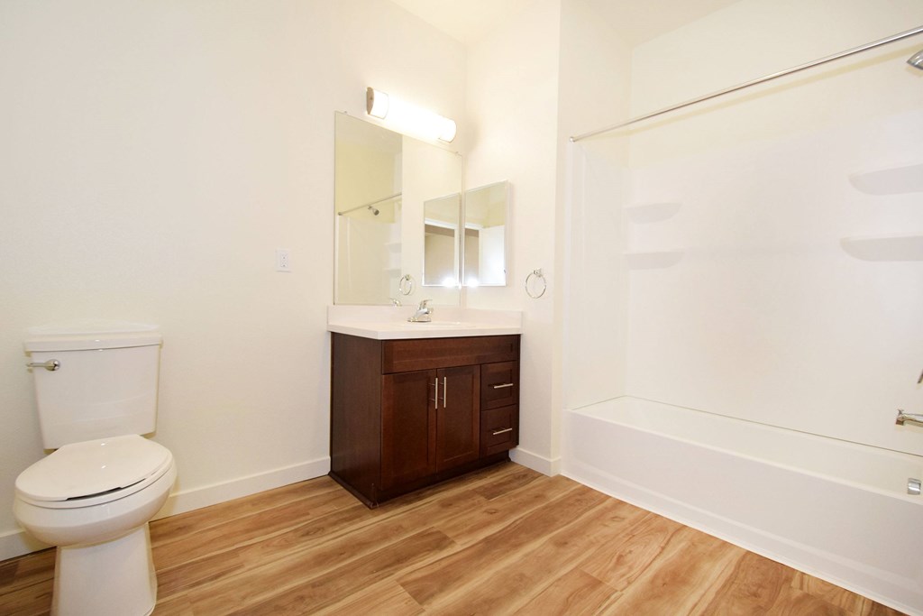 A white toilet sits in a bathroom with wooden floors and a white tub.