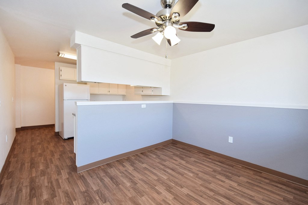 an empty living room with a ceiling fan and a kitchen in the background