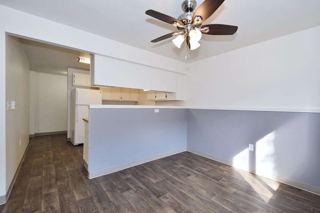 an empty living room with a ceiling fan and a kitchen in the background