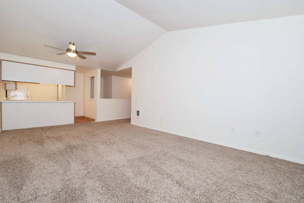 the spacious living room and kitchen of an apartment with carpeting and a ceiling fan