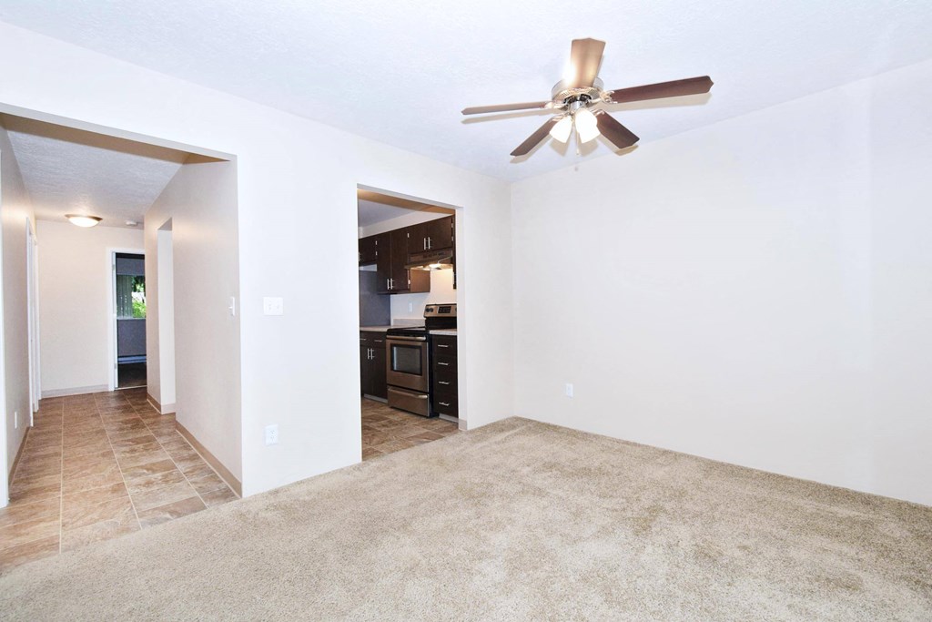 an empty living room with a ceiling fan and a kitchen in the background