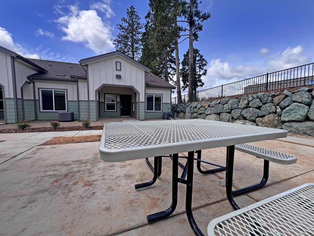 A picnic table is in the foreground of a building with a stone wall and trees in the background.
