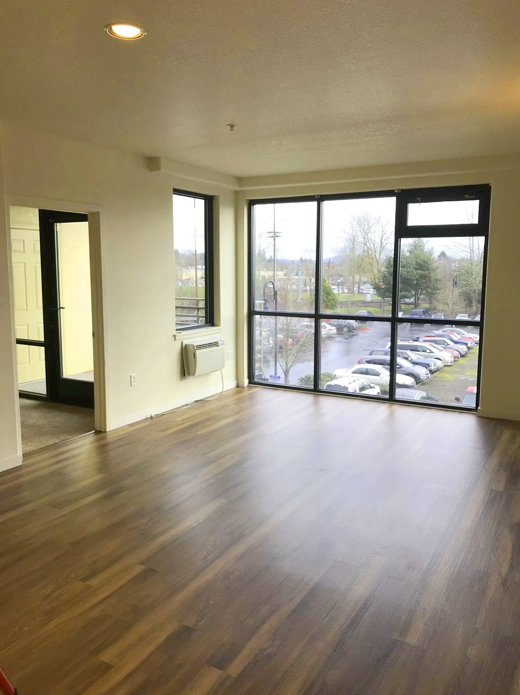 A room with wooden floors and a view of a parking lot through the windows.