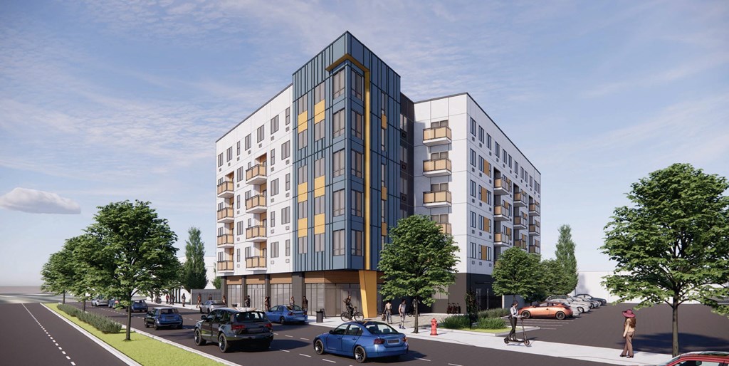 A modern multi-story building with a mix of white and blue panels is surrounded by trees and a street with cars and pedestrians.