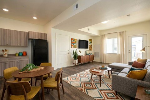 A living room with a grey couch, a wooden table and chairs, a black fridge, a flat screen TV, and a rug.