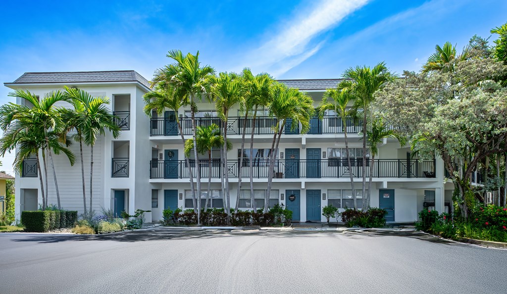 A white apartment building with blue doors and windows is surrounded by palm trees.