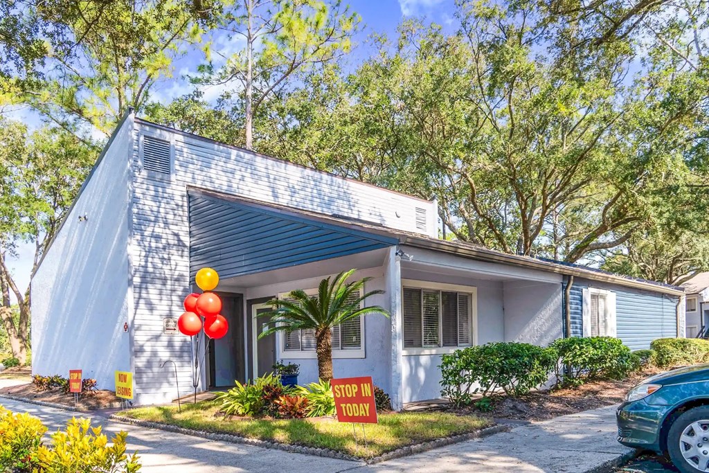 A house with a blue roof and a sign that says "STOP IN TODAY" is surrounded by trees and has a car parked in front.