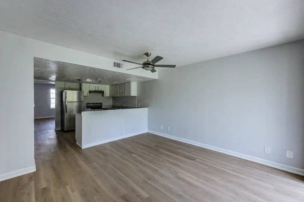 A room with a ceiling fan and a kitchen area in the background.