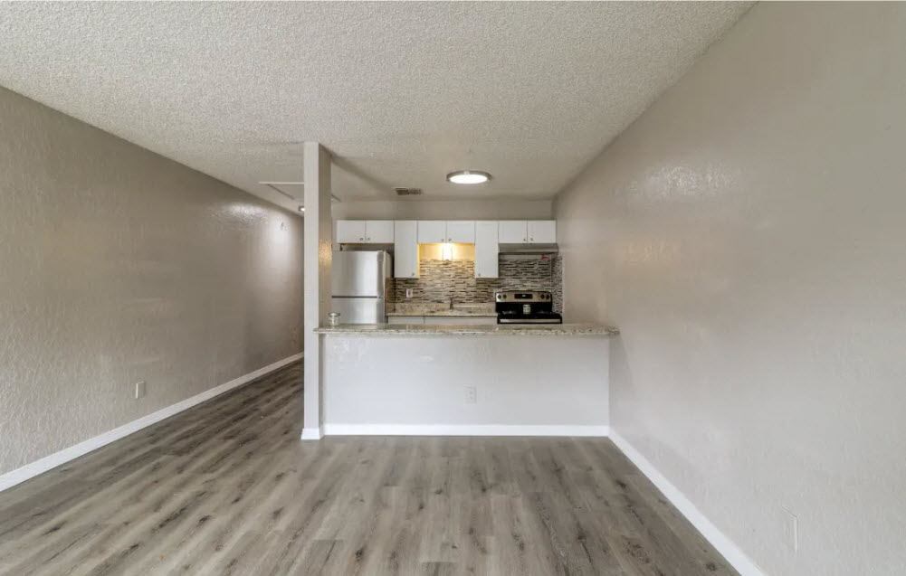 an empty living room with a kitchen in the background