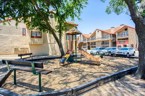 A playground with a slide, swings, and benches is surrounded by a fence and a building in the background.