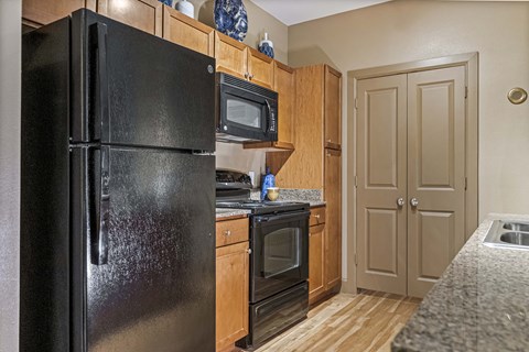 A black refrigerator stands in a kitchen with wooden cabinets and a microwave above the stove.