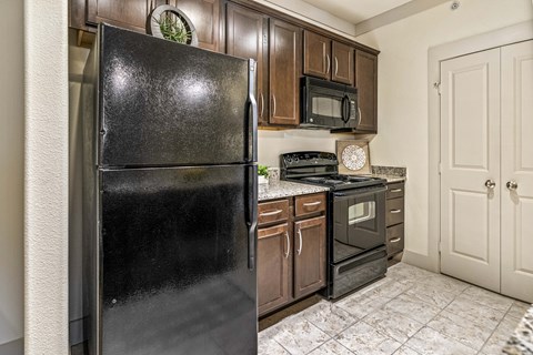 A black refrigerator stands in a kitchen with wooden cabinets and a tiled floor.