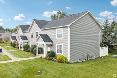 A house with a grey siding and a white door is surrounded by a green lawn.