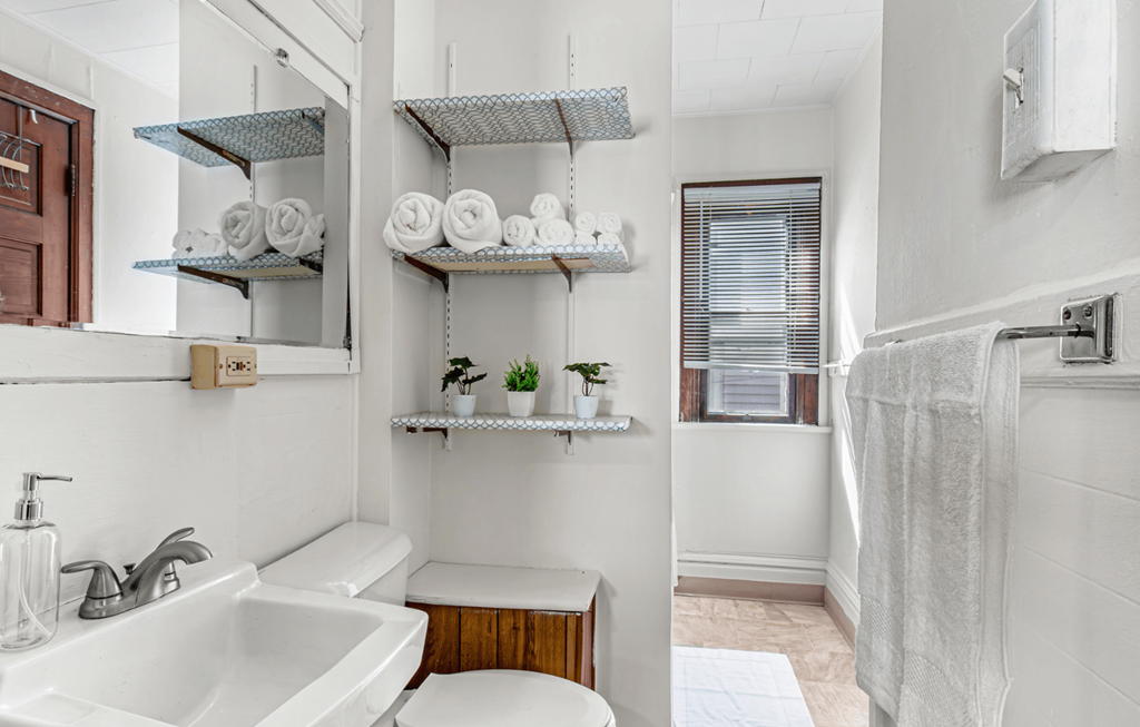 A white bathroom with a sink, toilet, and shelves with towels and plants.