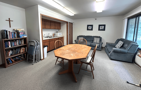 A living room with a wooden table and chairs, a bookshelf, and a kitchen area in the background.