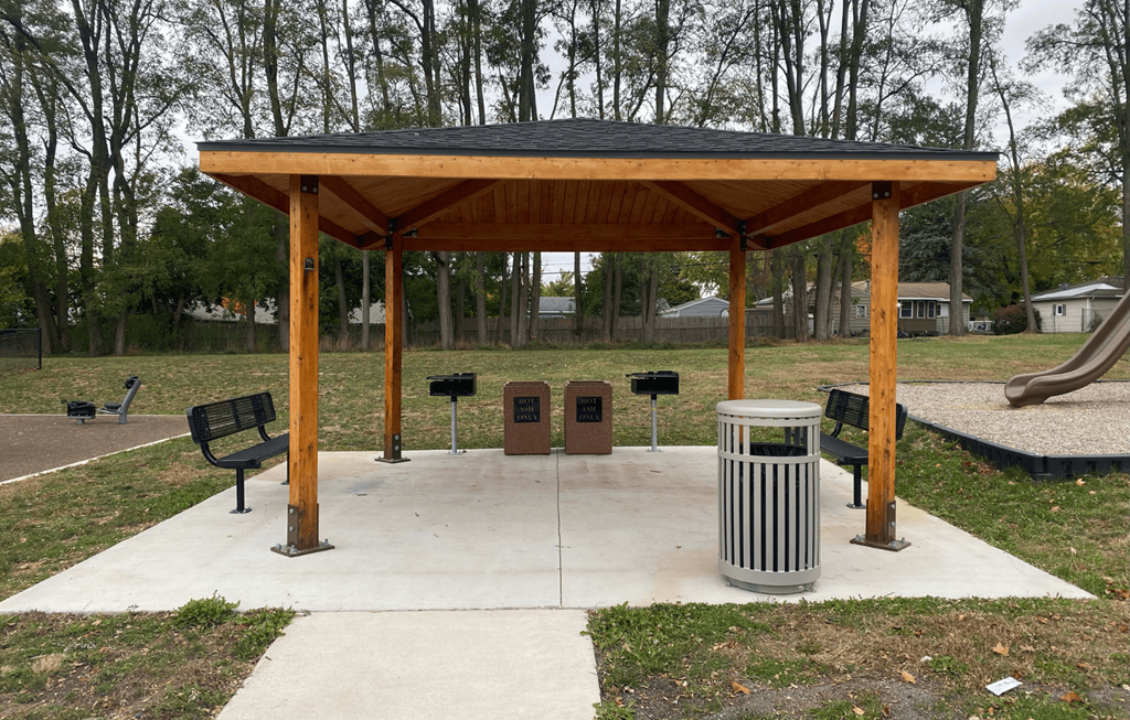 A wooden pavilion with a black roof is surrounded by a concrete walkway and a metal trash can.