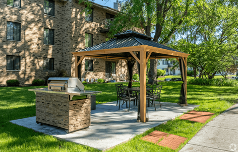 A wooden gazebo with a grill is situated in a grassy area in front of a building.