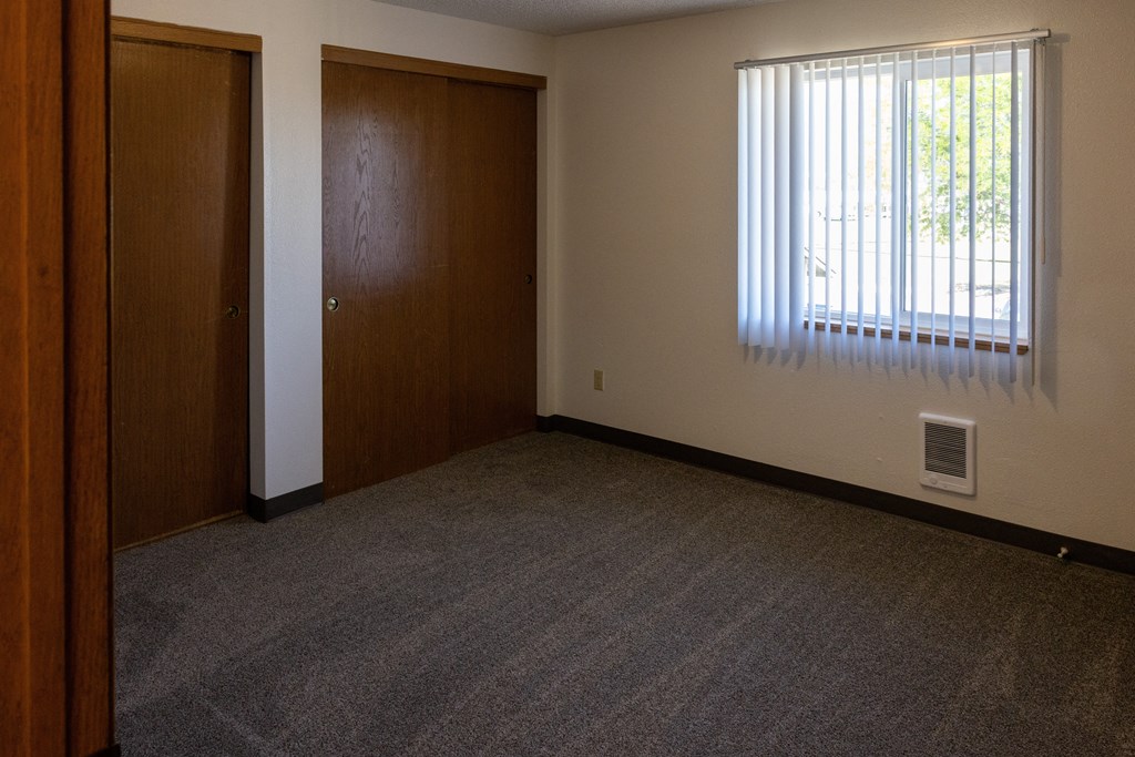 A room with a carpeted floor, a window with blinds, and two doors.