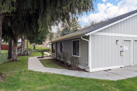 A small building with a grey roof and a white door is surrounded by a grassy area and trees.