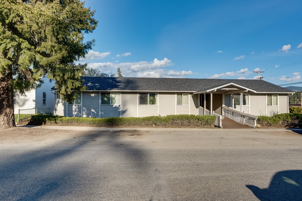 A house with a grey roof and white walls is surrounded by a driveway and a tree.