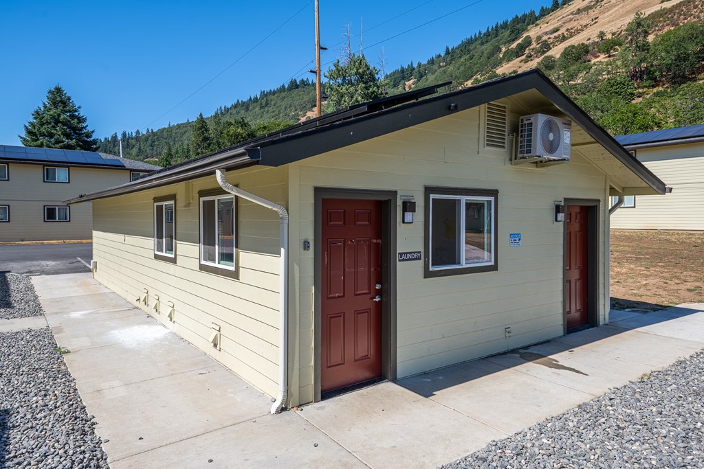 A small house with a brown door and a red door is surrounded by a gravel driveway.