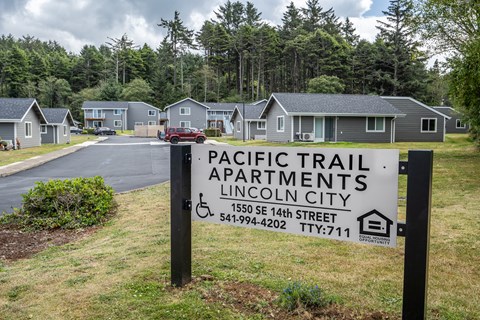 A sign for Pacific Trail Apartments in Lincoln City is displayed in front of a row of houses.