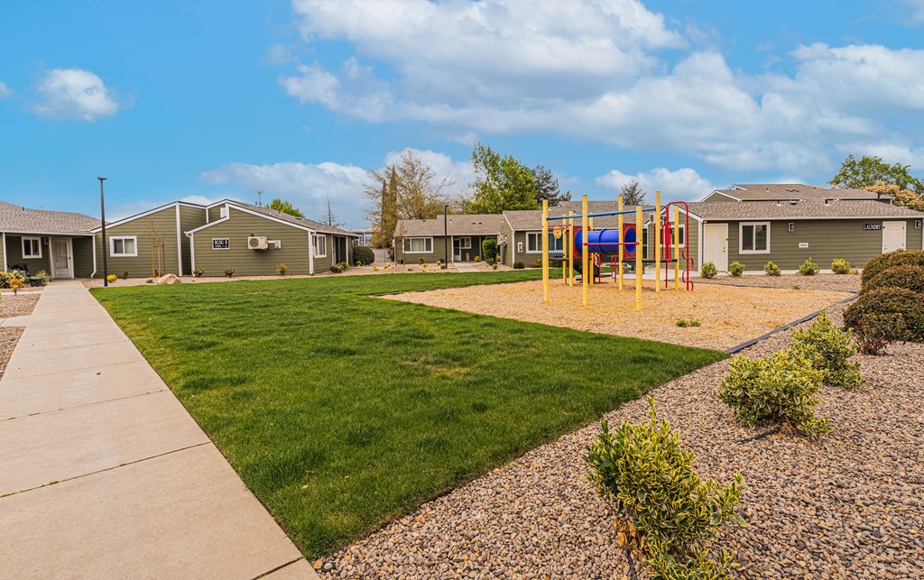 A playground with a yellow swing set is in the foreground of a residential area with houses and green grass.