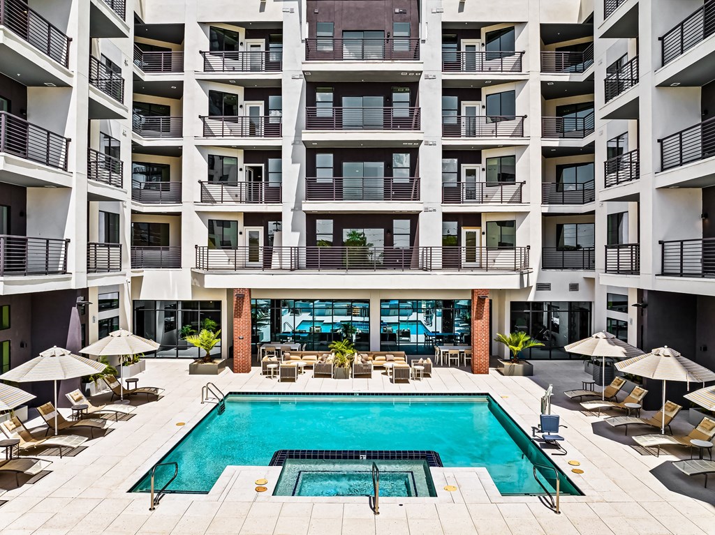 A large swimming pool in the middle of a courtyard surrounded by chairs and umbrellas.